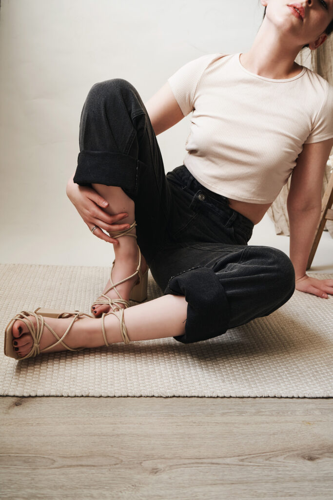 Gabriela Inzunza for Avice a relaxed pose on a textured rug wearing a white crop top and dark jeans with strappy sandals.